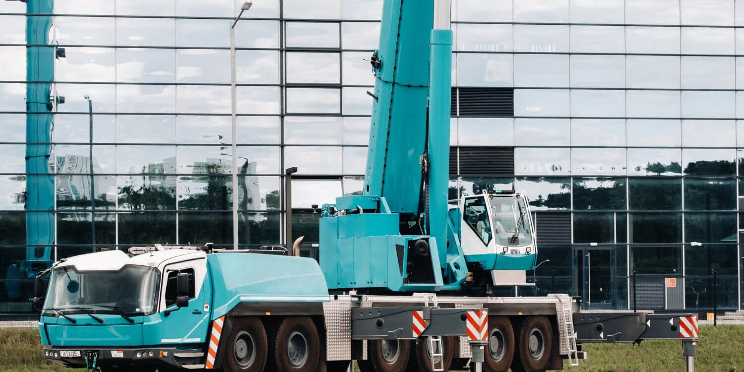 a-large-blue-truck-crane-stands-ready-to-operate-on-hydraulic-supports-on-a-platform-next-to-a-large-modern-building-the-largest-truck-crane-for-solving-complex-tasks